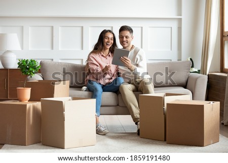 In cozy living room heap of carton boxes, couple sit on sofa with tablet buying furniture on internet, search design ideas, enjoy rest at new home. E-commerce house services, first flat owners concept Royalty-Free Stock Photo #1859191480