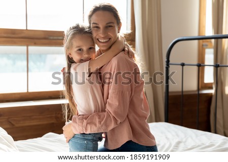In the morning woke up loving mother and little 6s sweet daughter embracing seated on comfy bed in bedroom smiling looking at camera. Family bonds, sincere love, happy motherhood, upbringing concept