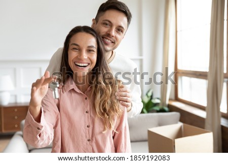 Happy spouses portrait, people standing in living room on relocation day smiling looking at camera, husband hug wife holding keys to the new house. Moving day, bank lending for young family concept