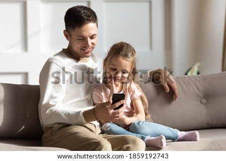 Dad and daughter sit on sofa in living room with smartphone looking at screen watching videos on internet, making videocall, using new application, having fun enjoy modern technologies at home concept