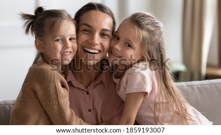 Portrait of happy family, mother day celebration concept. Single loving mommy and two little 6s daughters embracing seated on sofa at home. Young cheerful mom with cute girls smile looking at camera