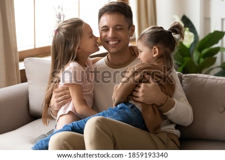 Cheerful laughing daddy having fun hugging two preschool 6s 7s daughters, kids seated on dad laps feels happy spend time together. Family bond and connection, father day holiday celebration concept