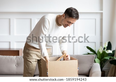 Man standing in living room on day of move to rented home, unbox carton box with belongings. Relocate to new own apartment collect personal things. Delivered parcel to satisfied client, tenant concept Royalty-Free Stock Photo #1859194243
