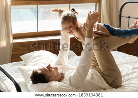 Lying on comfortable bed father raised his little daughter holding her hands, she seems flying in air. Dad and kid play game in morning, dream future travel together, spend free time have fun at home