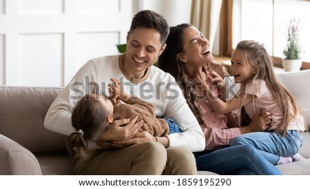 Funny parents play with two daughters resting on couch in light cozy living room. Family enjoy playtime together at home, tickling each other laughing feels happy. Having fun at home with kids concept Royalty-Free Stock Photo #1859195290