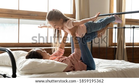 In the morning woman lies in bed play with 6s daughter, hold cute kid, girl stretched arms like airplane wings flying in air have fun with mom. Free time, games with child, dreams about travel concept