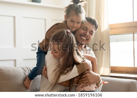 Two cute little daughters cuddling loving father congratulate him Happy Father Day sit on sofa having fun on weekend playing active games laughing feels overjoyed. Family bonds, love, devotion concept Royalty-Free Stock Photo #1859196106