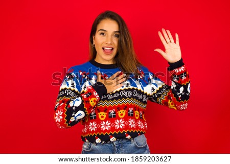 I swear, promise you not regret. Portrait of sincere Young beautiful Hispanic woman wearing winter sweater against red wall raising one arm and hold hand on heart as give oath, telling truth.