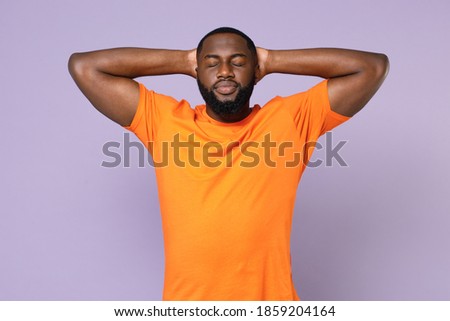 Relaxed young african american man 20s wearing basic casual blank orange t-shirt standing sleep with hands behind head keeping eyes closed isolated on pastel violet colour background studio portrait