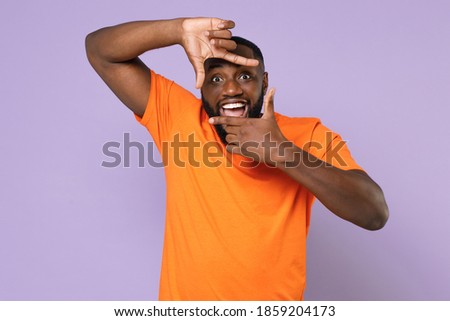 Excited young african american man 20s wearing basic casual blank orange t-shirt standing making hands photo frame gesture looking camera isolated on pastel violet colour background studio portrait