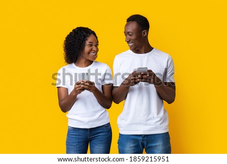 Smiling young african american man and woman playing together and competing in video games on smartphones, yellow studio background. Cheerful black couple playing online games together