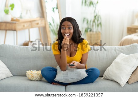 Full length portrait of young black woman with remote control watching TV and eating popcorn on sofa at home. Positive African American lady enjoying good movie or program on weekend Royalty-Free Stock Photo #1859221357