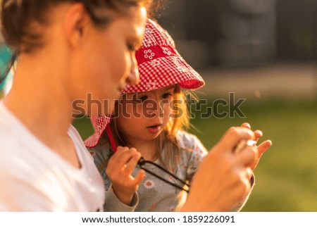 Mother shows little girl how to use digital camera during summer sunset. Child development concept.