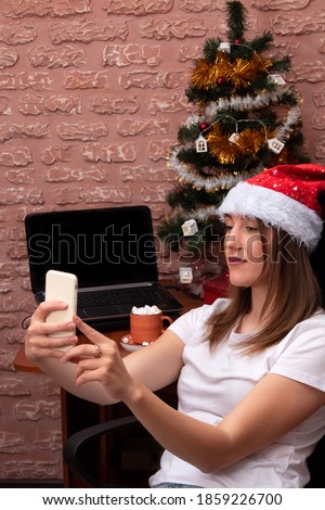 A girl takes a photo on her smartphone in a new year's mood. Table with laptop, Christmas tree, decorations.