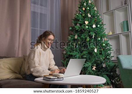 Happy smiling woman having online training course on laptop computer while sitting in home interior near beautiful decorated Christmas tree during winter holidays 