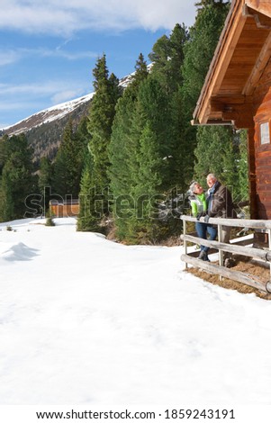 A smiling senior couple standing at the wooden fence outside the cabin in the snowy woods