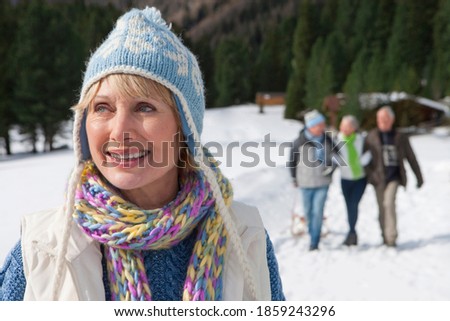 A close up of a smiling senior-adult woman standing in the snowy woods with her friends in the background