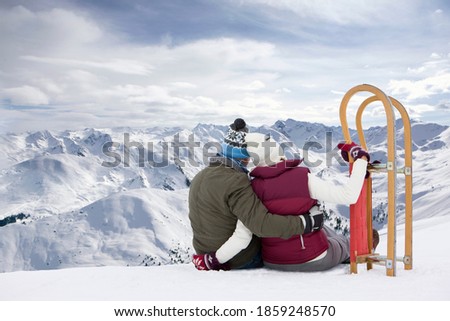 A romantic senior couple sitting and hugging at the top of a snowy mountain next to a sled