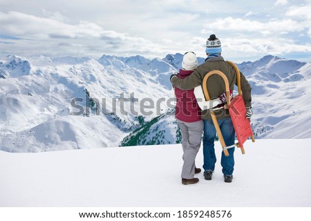 A romantic senior couple carrying a sled and standing at the top of a snowy mountain close to each other while enjoying the beautiful view