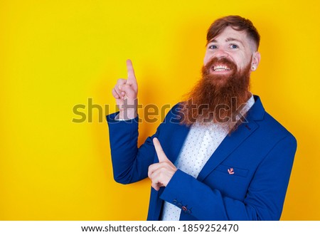 young red haired bearded businessman standing against yellow background indicating finger empty space showing best low prices, looking at the camera