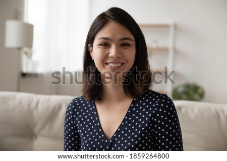 Head shot portrait smiling Asian young woman looking at camera, sitting on couch, attractive female blogger shooting vlog for social network, teacher coach recording webinar, working at home