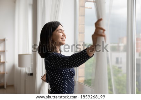 Smiling Asian young woman opening curtains in morning, enjoying sunlight, attractive female standing near window at home or in hotel, starting new day, dreaming, planning good future, visualizing Royalty-Free Stock Photo #1859268199