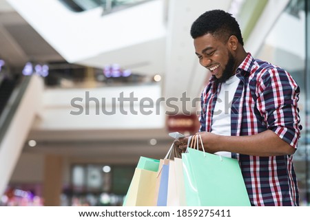 Cashback Bonus. Happy African Man Holding Shopping Bags And Smartphone In Mall, Using Discount App, Looking At Phone Screen And Smiling, Enjoying Online Offers, Standing In Modern Department Store Royalty-Free Stock Photo #1859275411