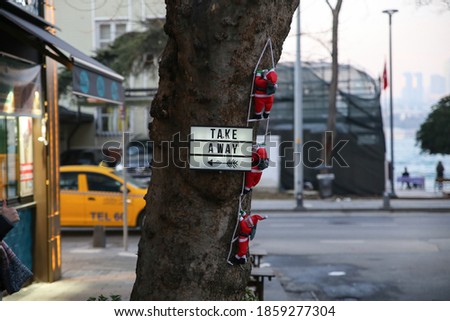 Next to a store, 3 toy santa clauses climbing onto the trunk of a brown tree with a yellow taxi behind them, and the word "take away" on the same tree.