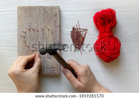 Step by step instruction: DIY gift for Valentine's Day. step 2: hands hammer nails into a wooden board in the form of a heart. red threads and nails lie nearby.