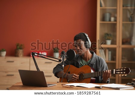 Wide angle portrait of talented African-American man singing to microphone and playing guitar while recording music in studio, copy space