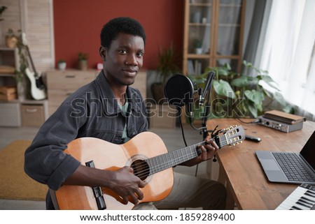 Portrait of young African-American man playing guitar and looking at camera while sitting by microphone in home recording studio, copy space
