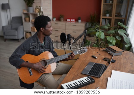 High angle portrait of young African-American man playing guitar and singing to microphone in home recording studio, copy space
