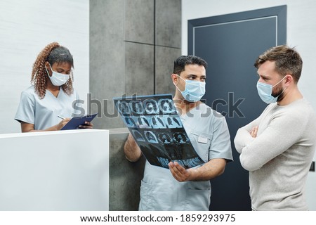 Serious mixed-race male radiologist in uniform and mask showing x-ray image to bearded patient against reception counter and young nurse