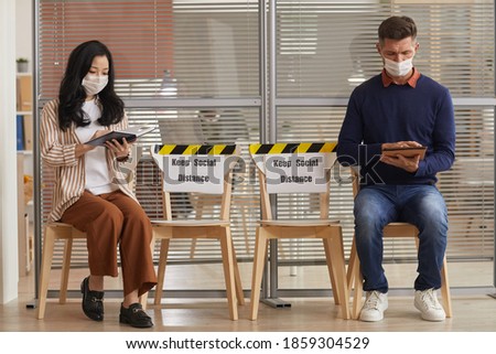 Full length portrait of young people wearing masks while waiting in line in office with Keep Social Distance signs, copy space