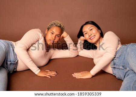 Two young contemporary females of various ethnicities in blue jeans and white pullovers looking at you with toothy smiles while lying on the floor