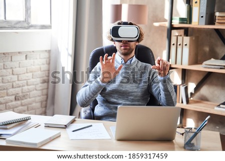 Excited young man wearing virtual reality glasses sitting at table with laptop, while reading and scrolling something at the virtual screen. Stock photo