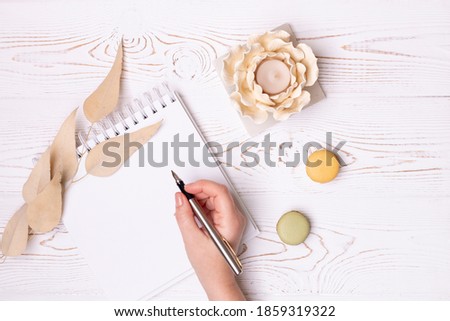 A female hand holds a pen over a clean diary with spring, macaroons, beige leaves and a flower-shaped candlestick on a white shabby wooden table. Fashion flat lay