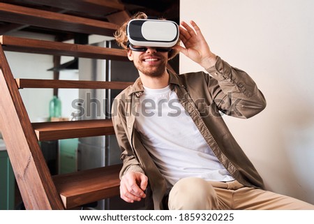 Handsome young caucasian man adjusting his VR headset with toothy smile while sitting at the stairs at the cozy living room. Stock photo