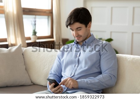 Pleasant conversation. Happy confident millennial man resting on sofa at living room on weekend using smartphone texting message email to beloved woman, reading good news from friend, chatting online