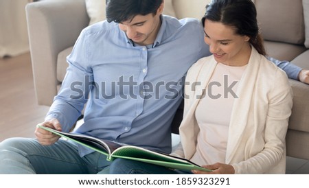 Pleasant pastime. Happy millennial spouses newlyweds sitting close together on warm floor at home looking over interior design catalogue choosing furniture stylish decor for new house flat apartment
