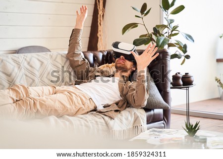 Testing new technologies. Attractive young unshaven man in VR headset gesturing and smiling while laying at the sofa at home. Stock photo
