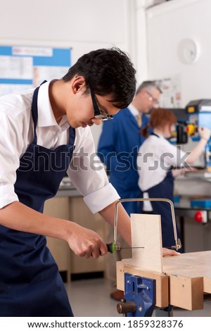 A portrait shot of a young boy with glasses sawing wood in a vise machine in a classroom.