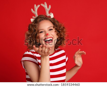delighted funny young woman in a Christmas reindeer costume laughs and blows a kiss on colorful red background
