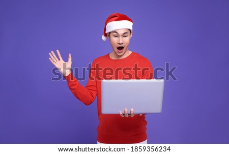 Young asian man in red casual attire wearing Santa hat and holding laptop and looking at screen excited face and surprise hand on purple background.Happy new year concept.