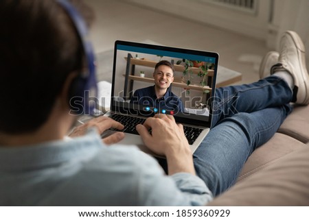 Rear shoulder view relaxed young man in earphones holding video call online web meeting with caucasian male friend or colleague, using computer modern distant communication application at home. Royalty-Free Stock Photo #1859360929
