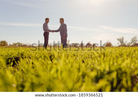 Full body side view of male farm owner and agronomist standing in middle of green grassy field and discussing professional issues in summer day in countryside Royalty-Free Stock Photo #1859366521