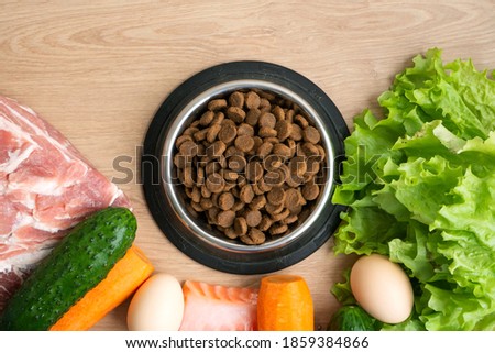 Dry pet dog food with natural ingredients. Raw meat, fish, vegetables, eggs and salad near bowl with dry pet feed on wooden background. concept of a correct balanced and healthy nutrition for pet