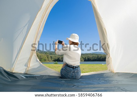 Young Asian woman in a T-shirt, shorts, and a bucket hat sitting in a camping tent taking a picture of a beautiful calm lake with dense woods and clear blue sky with her smartphone on a warm sunny day