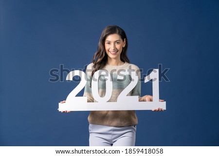 Happy young Asian woman holding 2021 white paper banner for celebrate merry Christmas and happy new year on blue color background, celebration and decoration for holiday event concept