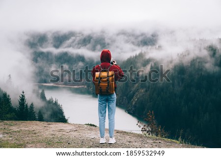 Back view of anonymous person with orange rucksack photographing nature on misty day while spending time on rocky area in nature